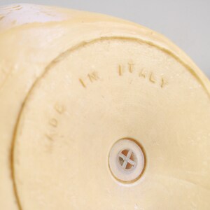 May include: Close-up of the bottom of a cream-colored ceramic figurine. The figurine has a small hole in the bottom and the words "MADE IN ITALY" are embossed on the bottom.