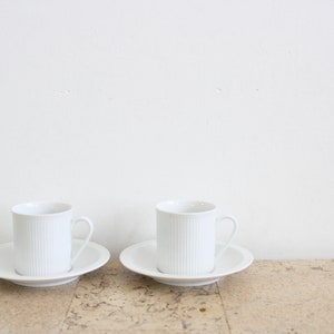 May include: Two white ceramic coffee cups with saucers. The cups have a ribbed design and are placed on a light brown surface.