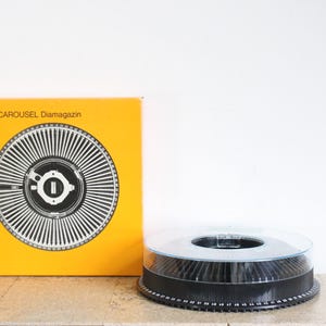 May include: A yellow box with the text "KODAK CAROUSEL Diamagazin" and a black and white image of a slide carousel. A black slide carousel with a clear plastic cover is sitting on a light brown surface.