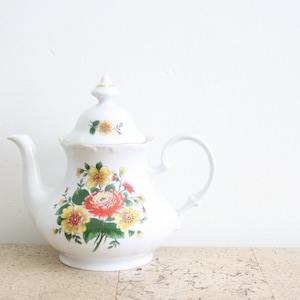 Puede incluir: Tetera de porcelana blanca con un diseño floral con flores rojas, amarillas y verdes. La tetera tiene una tapa y un asa.