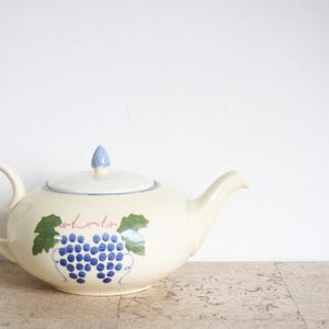Könnte beinhalten: Cremefarbene Keramik-Teekanne mit Deckel und Ausguss. Die Teekanne ist mit einem blauen Traubenmuster und grünen Blättern verziert. Der Deckel hat einen blauen Knauf. Der Griff hat eine Schlaufenform.