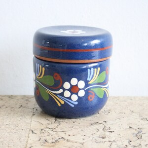 May include: A blue ceramic jar with a lid, decorated with white flowers and green leaves. The jar has a brown band around the top.