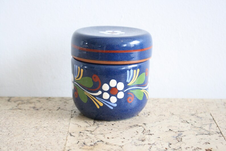 May include: A blue ceramic jar with a lid, decorated with white flowers and green leaves. The jar has a brown band around the top.