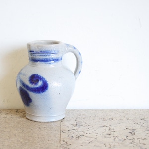 May include: A small, off-white ceramic pitcher with a blue handle and decorative blue swirls. The pitcher has a rounded body and a narrow neck, with blue bands around the rim. The pitcher is likely vintage or antique.