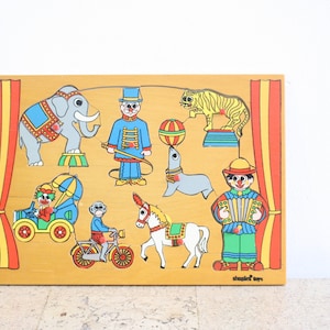 Puede incluir: Un rompecabezas de circo de madera con personajes y animales coloridos. El rompecabezas incluye un payaso, un elefante, un tigre, una foca, un mono, un caballo y un coche. El rompecabezas está hecho por Schlegel Toys.