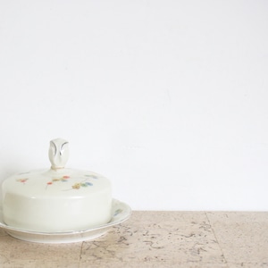 Peut inclure: Un beurrier en céramique crème avec un couvercle et une petite poignée. Le couvercle est décoré de petits motifs floraux rouges, bleus et jaunes. Le plat repose sur une assiette assortie avec un bord festonné.