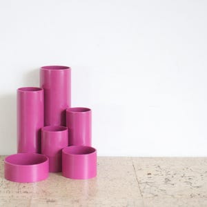 Puede incluir: Colección de seis objetos cilíndricos de color magenta, de diferentes alturas y diámetros. Los objetos están dispuestos en grupo, con el más alto en la parte trasera. Los objetos son de material liso, sobre fondo blanco. Medidas en centímetros.