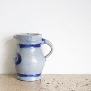 May include: A stoneware pitcher with a light gray body and blue accents. The pitcher has a handle and a spout. Blue bands encircle the top and bottom, with a decorative swirl on the side. A classic piece for serving beverages.