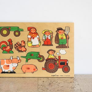 Könnte beinhalten: Ein rechteckiges Holz-Steckpuzzle mit farbenfrohen Illustrationen zum Thema Bauernhof, darunter ein grüner Traktor, ein Hahn, ein Huhn, eine Kuh, ein Schwein, eine Schubkarre, ein roter Traktor und drei Figuren mit roten Holzknöpfen.