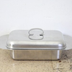 Puede incluir: Una mantequera rectangular de acero inoxidable con tapa y asa. El plato tiene un dise&ntilde;o sencillo y funcional con una base ligeramente ensanchada y una tapa a juego. El asa tiene forma de barra curva.
