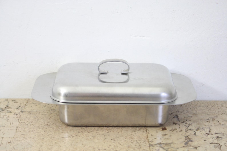 Puede incluir: Una mantequera rectangular de acero inoxidable con tapa y asa. El plato tiene un dise&ntilde;o sencillo y funcional con una base ligeramente ensanchada y una tapa a juego. El asa tiene forma de barra curva.