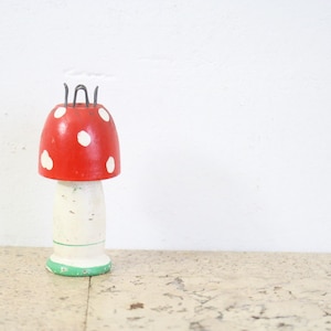 May include: A whimsical, hand-painted mushroom-shaped object. The mushroom cap is bright red with white polka dots. The stem is off-white, and the base is green. Metal wire prongs extend from the top. A decorative item.