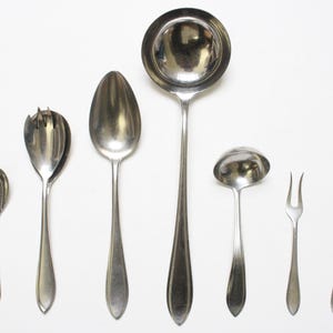 May include: An assortment of vintage silverware, including spoons, a ladle, a fork, and a knife, all with a shiny, metallic finish. The utensils are arranged on a white surface, showcasing their classic design and elegant shapes.