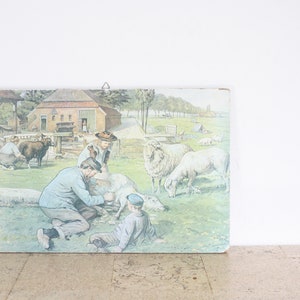 May include: Vintage framed print depicting a pastoral scene with people shearing sheep. The artwork features a barn, sheep, and figures in period clothing, rendered in soft colors. The print is mounted on a wooden board.