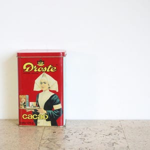 Può includere: Una scatola rettangolare rossa vintage di cacao Droste. La scatola presenta una donna con cuffia bianca e abito scuro che tiene un vassoio. La parola "cacao" è stampata in bianco e la scatola ha un logo dorato.