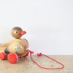 May include: A wooden pull-along duck toy with a brown head, red beak, and red wheels. The duck's body is light brown with blue and white painted details. A red string is attached for pulling.
