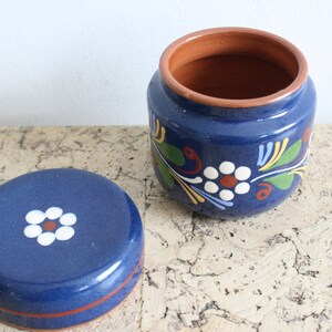 May include: A blue ceramic jar with a lid, both decorated with white flowers and green leaves. The jar has a brown rim.