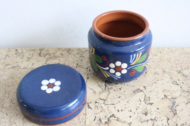 May include: A blue ceramic jar with a lid, both decorated with white flowers and green leaves. The jar has a brown rim.