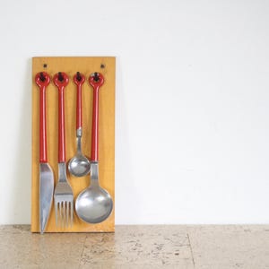 May include: A set of five silver utensils with red handles mounted on a wooden board. The utensils include a knife, fork, and three spoons.