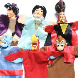 Könnte beinhalten: Fünf bunte Handpuppen mit Figuren aus dem Disney-Film Aladdin. Die Puppen sind Aladdin, Jasmin, Dschafar, Abu und der Dschinn. Die Puppen sind aus weichem Stoff gefertigt und haben detaillierte Gesichtszüge.
