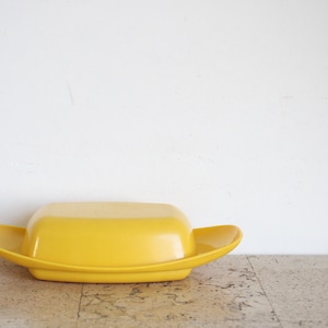 May include: A vibrant yellow butter dish with a lid, placed on a speckled surface. The dish features a rectangular base with curved, wing-like sides. The lid is rectangular and fits neatly on the base. A kitchenware item.