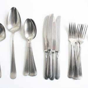 May include: An assortment of vintage silverware, including spoons, knives, and forks, arranged on a white surface. The silverware has a silver finish and a classic design, perfect for a formal dining setting.