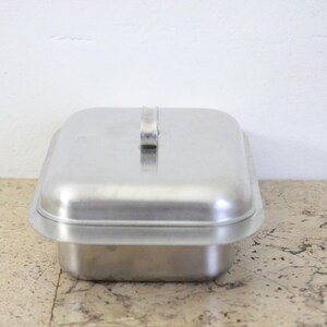 Puede incluir: Una fuente rectangular de metal cepillado con tapa a juego. La tapa tiene un asa peque&ntilde;a y rectangular. La fuente se encuentra sobre una superficie clara y moteada contra una pared blanca. La fuente probablemente se utiliza para cocinar o almacenar.