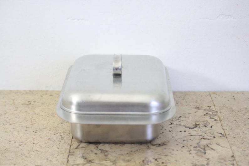 Puede incluir: Una fuente rectangular de metal cepillado con tapa a juego. La tapa tiene un asa peque&ntilde;a y rectangular. La fuente se encuentra sobre una superficie clara y moteada contra una pared blanca. La fuente probablemente se utiliza para cocinar o almacenar.