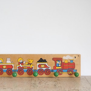 Puede incluir: Perchero de madera con temática de tren, con una combinación de colores rojo, azul y amarillo. Los vagones del tren presentan ilustraciones de niños, un gato y un oso de peluche. El perchero tiene pomos verdes para colgar objetos.