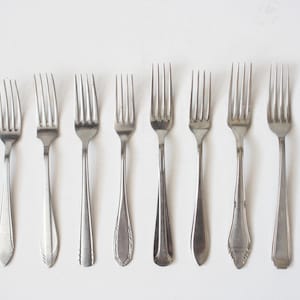 May include: A collection of ten silver-toned metal forks arranged in a horizontal row. Each fork features a distinct handle design, ranging from simple smooth finishes to detailed decorative patterns at the base of the stems and tines.