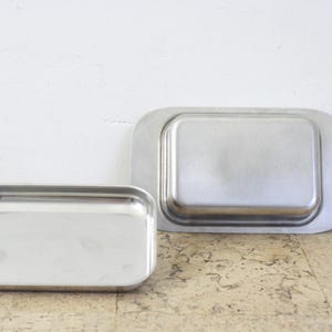 Puede incluir: Dos bandejas rectangulares de acero inoxidable. La bandeja m&aacute;s peque&ntilde;a est&aacute; a la izquierda, con un borde ligeramente redondeado. La bandeja m&aacute;s grande est&aacute; a la derecha, con un borde m&aacute;s redondeado. Ambas bandejas tienen un acabado met&aacute;lico brillante.