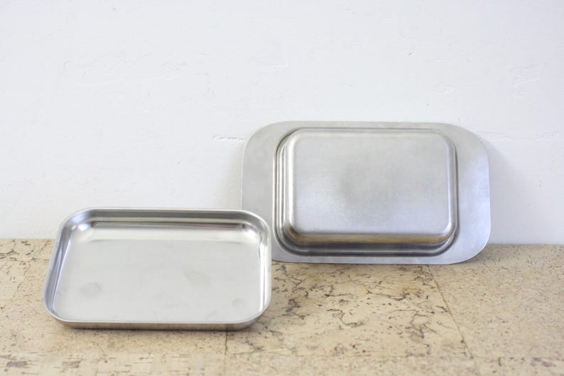 Puede incluir: Dos bandejas rectangulares de acero inoxidable. La bandeja m&aacute;s peque&ntilde;a est&aacute; a la izquierda, con un borde ligeramente redondeado. La bandeja m&aacute;s grande est&aacute; a la derecha, con un borde m&aacute;s redondeado. Ambas bandejas tienen un acabado met&aacute;lico brillante.