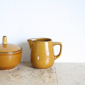 May include: A ceramic sugar bowl with a lid and a small creamer pitcher. Both are a warm, golden-brown color with a glossy finish and dark brown trim. The sugar bowl has a rounded shape, and the creamer has a handle and spout.