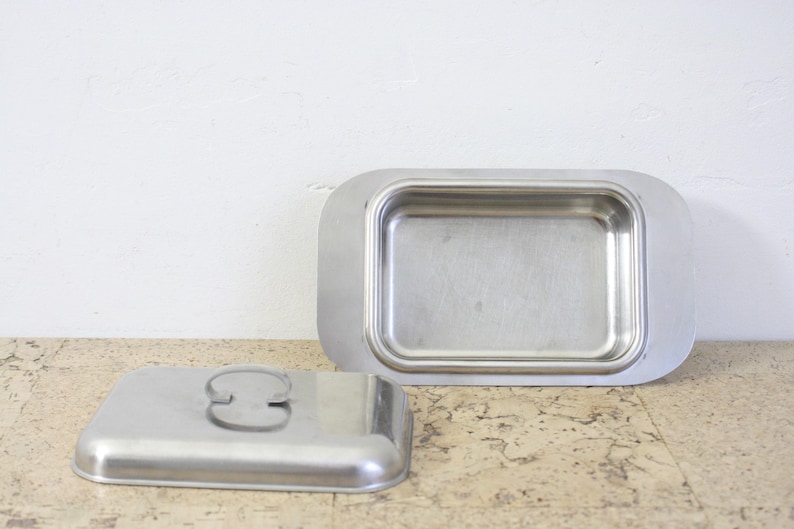 Puede incluir: Una mantequera rectangular de acero inoxidable con tapa. El plato tiene esquinas redondeadas y un asa en la tapa. La tapa est&aacute; a un lado, revelando el interior del plato.