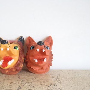 May include: Two dog-shaped masks. One has a tan face, red tongue, and green eyes, framed by brown fur. The other is reddish-brown with blue eyes and white teeth. Both have black noses. Ideal for fancy dress or theatre.