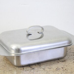 Puede incluir: Una fuente rectangular de acero inoxidable con tapa y asa a juego. La fuente tiene una base ligeramente ensanchada y una tapa redondeada con un asa curva. La fuente est&aacute; sobre una superficie clara.