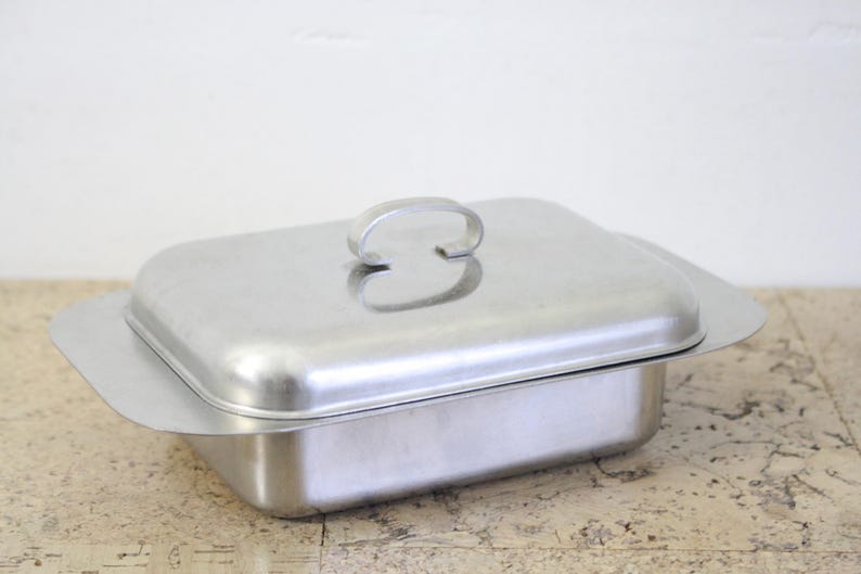 Puede incluir: Una fuente rectangular de acero inoxidable con tapa y asa a juego. La fuente tiene una base ligeramente ensanchada y una tapa redondeada con un asa curva. La fuente est&aacute; sobre una superficie clara.