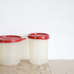 Puede incluir: Dos recipientes de plástico transparente con tapas rojas. Los recipientes son de diferentes tamaños y están sobre una superficie de corcho.