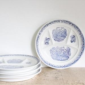 May include: A stack of four white ceramic divided plates featuring a blue transferware pattern. Each plate has three sections and is decorated with detailed blue illustrations of rural landscapes, trees, and houses, with a floral border.