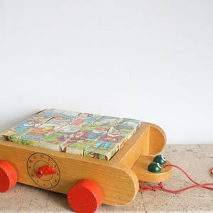 May include: Vintage wooden pull-along toy featuring a puzzle with colorful illustrations. The toy has a clock face, red wheels, and a pull string. The puzzle pieces depict cartoon characters. A classic children's toy.