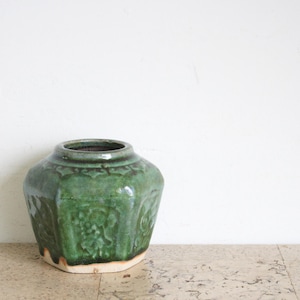 Może przedstawiać: Zielony ceramiczny wazon o szerokim korpusie i wąskim otworze. Wazon ma teksturowaną powierzchnię z wytłoczonymi wzorami kwiatowymi. Podstawa wazonu jest w jasnobeżowym kolorze. Wazon jest ustawiony na białym tle.