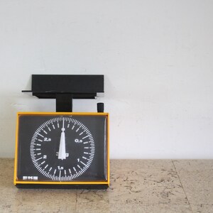 May include: A mechanical kitchen scale with a yellow frame and a black face. The circular dial features white markings and numbers ranging from 0.5 to 3 kg, with a central white needle pointing upwards. A black metal platform sits atop the scale.