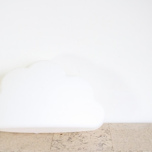 Könnte beinhalten: Eine weiße, wolkenförmige dekorative Leuchte. Das Licht besteht aus einem glatten, undurchsichtigen Material und steht auf einer strukturierten Oberfläche vor einer weißen Wand. Die Leuchte ist als flauschige Wolke gestaltet.