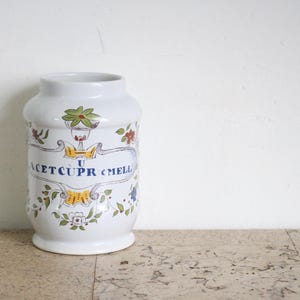 Può includere: Vaso in ceramica bianca con disegni floreali e banner blu, gialli e rossi. Il vaso ha una base larga e un collo leggermente più stretto. Il testo "ACETCU PR CMELL" è stampato sul vaso.