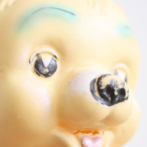 May include: Close-up of a yellow plastic toy bear with black painted eyes, a black painted nose, and a pink painted tongue. The bear has a blue painted eyebrow.