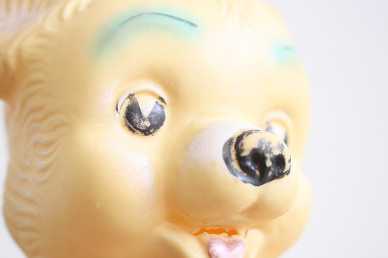 May include: Close-up of a yellow plastic toy bear with black painted eyes, a black painted nose, and a pink painted tongue. The bear has a blue painted eyebrow.