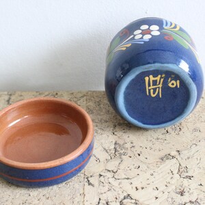 May include: A blue ceramic jar with a floral design and a brown ceramic lid. The bottom of the jar has the text "M'oi" in gold.