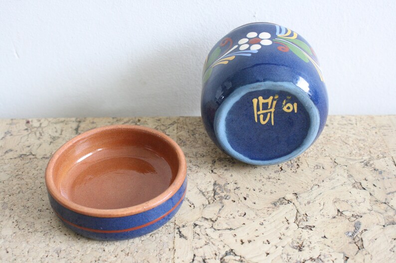 May include: A blue ceramic jar with a floral design and a brown ceramic lid. The bottom of the jar has the text "M'oi" in gold.