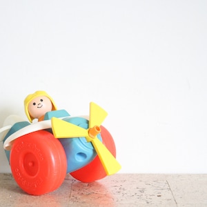 May include: A vintage toy airplane with a yellow propeller, blue body, and red wheels. A smiling figure with yellow hair sits in the cockpit.