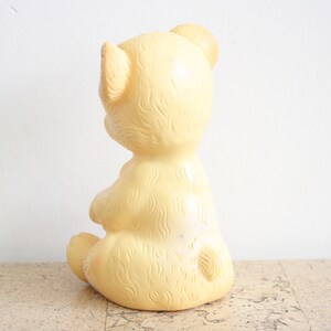 May include: A yellow rubber teddy bear toy sitting on a cork surface. The bear has a simple design with a smooth, rounded body and a small, pointed nose.